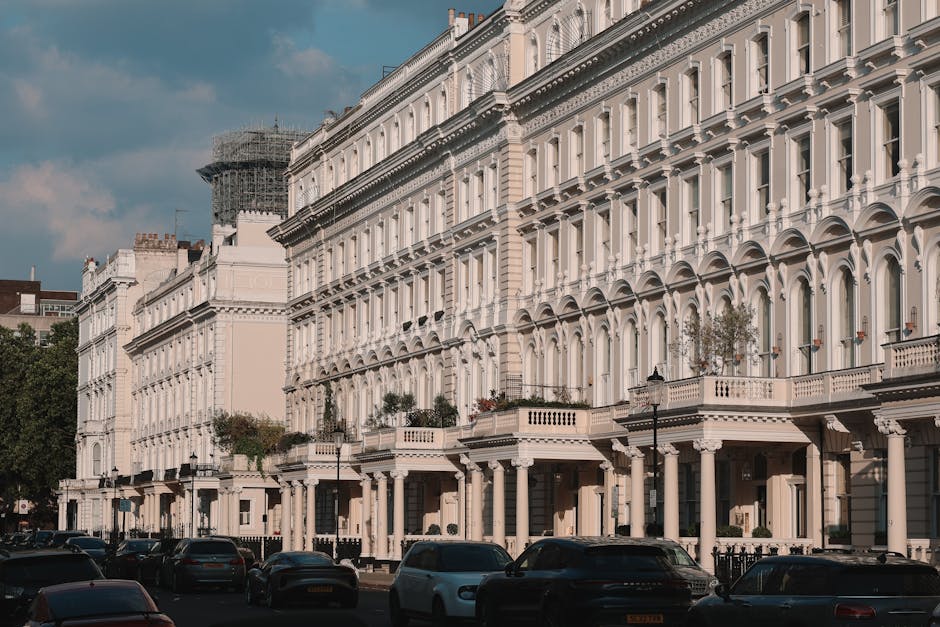 A row of elegant, white Victorian-style townhouses with detailed facades featuring large arched windows, decorative cornices, and balconies with railings. The buildings are uniformly aligned along a s