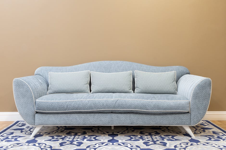 A light blue upholstered sofa with three matching patterned cushions is positioned against a beige wall in a living room. The sofa has rounded armrests and white wooden legs, sitting atop a decorative