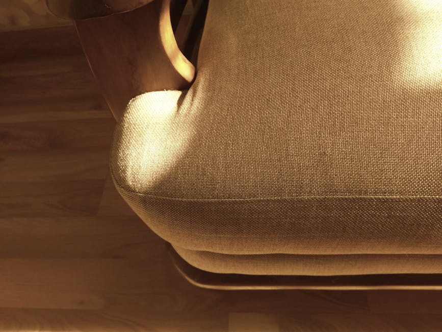 A light blue upholstered sofa with three matching patterned cushions is positioned against a beige wall in a living room. The sofa has rounded armrests and white wooden legs, sitting atop a decorative