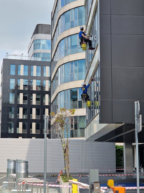 Close-up view of the exterior of a modern multi-story building during daytime, with workers cleaning the large glass windows using squeegees and safety harnesses. The building features a combination o